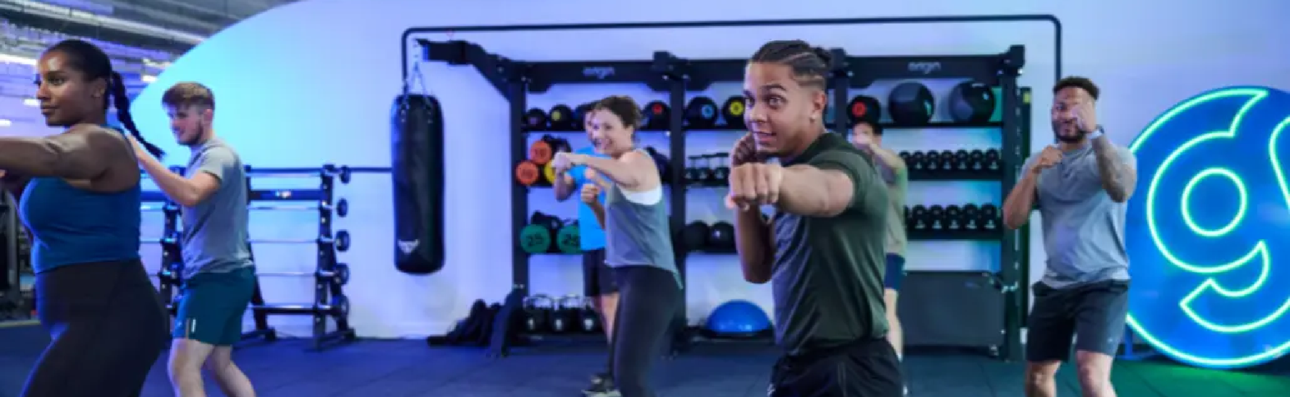 Group fitness class participants exercising in a modern gym with blue neon lighting and weight racks along the wall