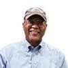 A headshot of a man of colour, presumably in his sixties, wearing a light blue button down shirt, glasses and a ball cap.